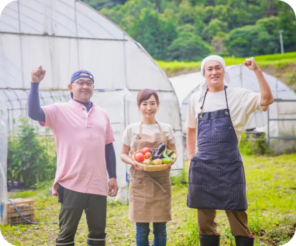 ピンクの服を着た男性と野菜を持って笑顔の女性と、エプロンをつけている男性の写真